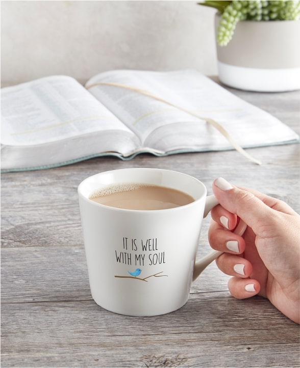 open bible and plant on tabletop, hand holding coffee mug printed with IT IS WELL WITH MY SOUL